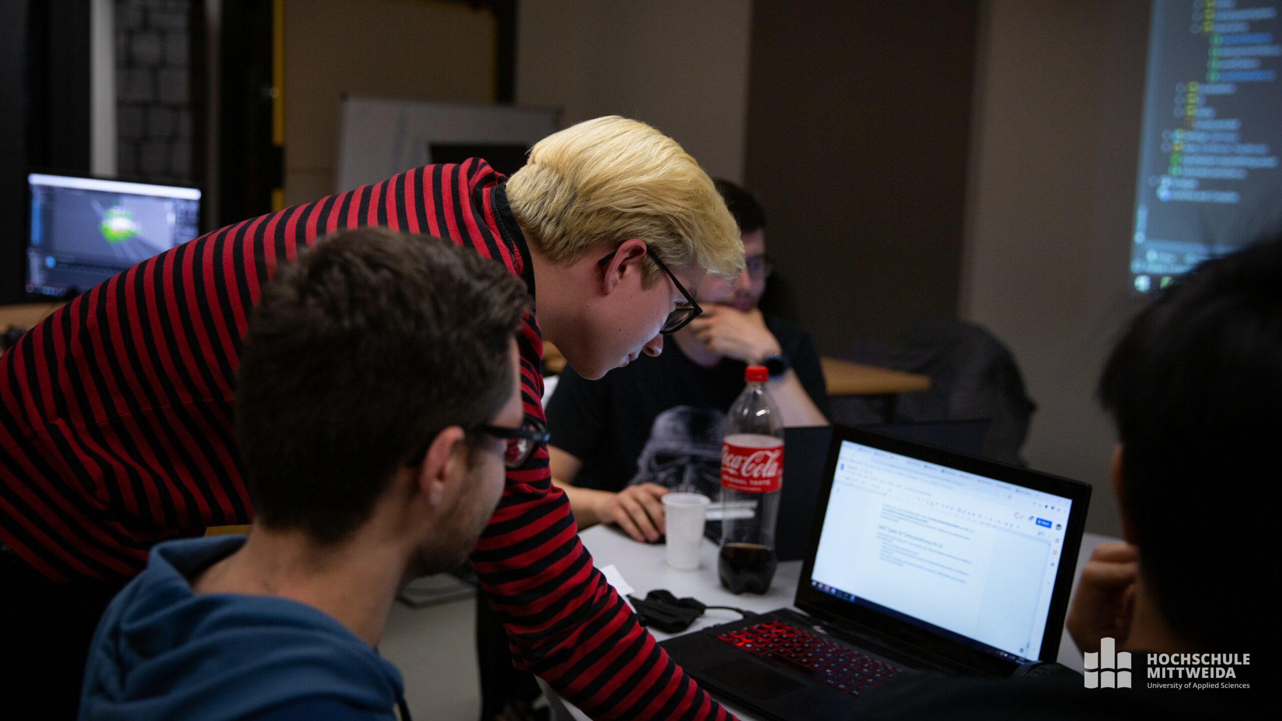 Drei Männer sitzen an einem Tisch, ein Mann im rotgestreiften Shirt arbeitet am Laptop.